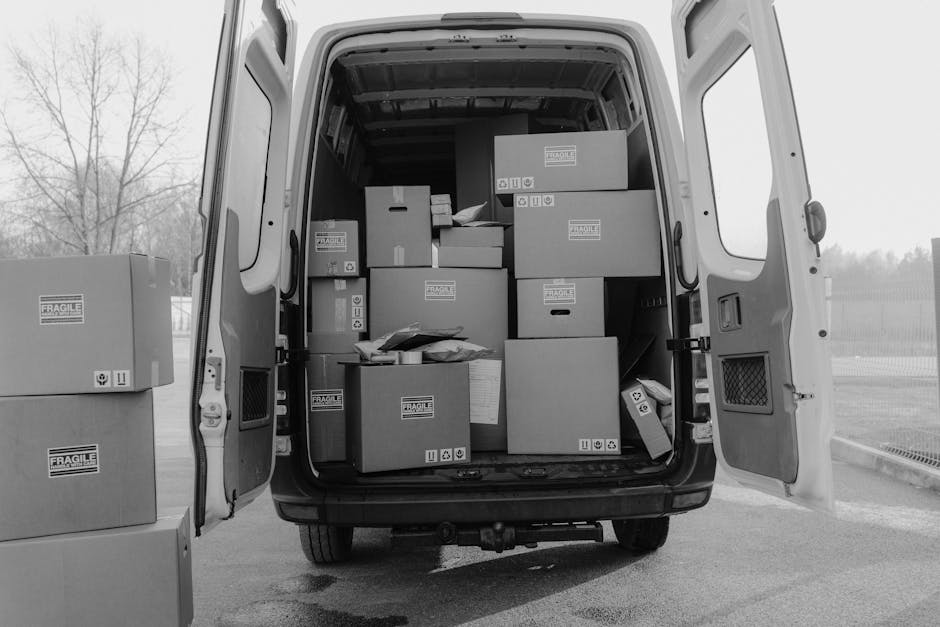 Inside the open rear of a white delivery van, four large cardboard boxes are stacked neatly, with three positioned on the floor and one resting on top of two others. The boxes are plain brown, with so